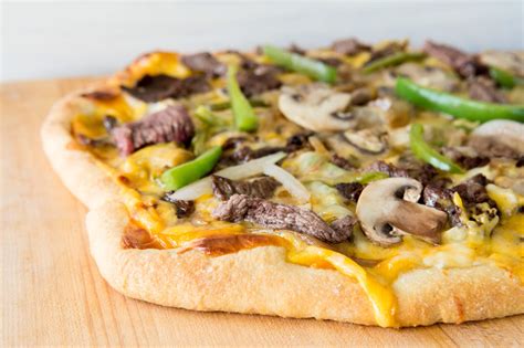 Cheese steak pizza. Set racks in the top and bottom thirds of the oven and preheat to 500 degrees. Roll out one dough half into a 14-inch oval on a board that has been gently dusted with flour. Place the dough on a baking sheet covered with parchment paper and brush with 1 teaspoon of olive oil. Repeat with the second half of the dough. 