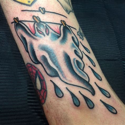 This arm tattoo features a figure with a hoodie, wearing a frightening ghost mask. The mask has big black eye sockets and a wide-open mouth, both inked with black color. The inverted heart-shaped nose is also done in black ink. The figure’s hands are visible, with a knife in the left hand and a Korean heart gesture with the right hand.. 