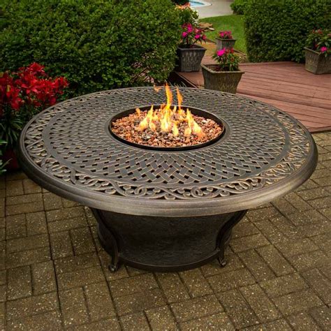 A ‘Patio Glow’ contemporary propane fire pit, with a metal frame. The central bowl is filled with rocks (included) which give off a smoldering glow when the gas jets are lit. There is an octagonal shelf around the central bowl, with removable ceramic tiles. It has a square base with tan synthetic fabric mesh panels, with the propane tank .... 